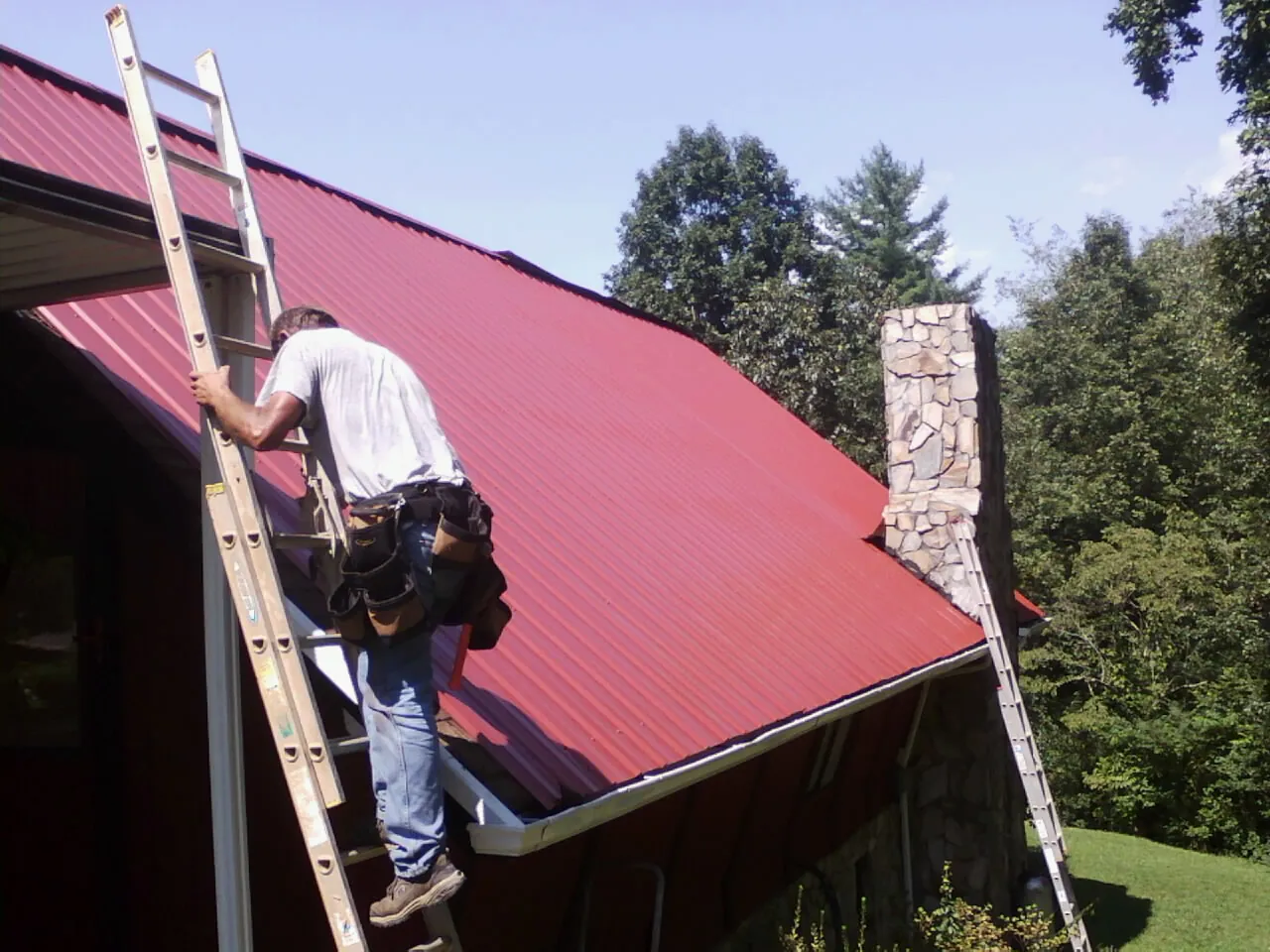 Roofer on a ladder beside a red metal roof and stone chimney — Soffit & Fascia Repair by Roofing Brothers in Baltic